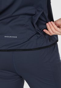 Marineblå atletisk top med en lynlåslomme i siden, der har en glat tekstur og en subtil logo detalje nær kanten.