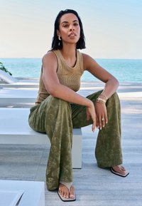 Débardeur côtelé de couleur tan associé à un pantalon large vert à motifs et des sandales plates. Bijoux simples pour accentuer, assis devant un paysage côtier.