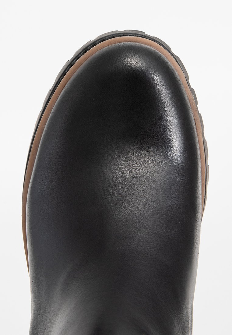 Zbliżenie na czubek czarnej skórzanej boty z brązowym otokiem i grubą, teksturowaną czarną podeszwą na białym tle.