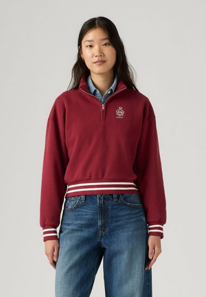 Junge Frau trägt ein bordeauxfarbenes Sweatshirt mit halbem Reißverschluss und gestreiften Bündchen und Saum über einem blauen Hemd und blauer Jeans, stehend vor einem einfarbigen Hintergrund.