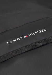 Texture nera con strisce verticali, con un logo bianco "TOMMY HILFIGER" in evidenza. Il design include un accento rettangolare in rosso e blu.