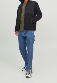 Zwarte fleecejas met een contrasterend nylon bovendeel, olijfgroen t-shirt, blauwe spijkerbroek en witte sneakers met zwarte accenten.