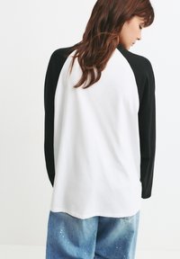 Langærmet T-shirt med en hvid krop og sorte raglanærmer. Har en rund halsudskæring, blødt stof og en afslappet pasform.