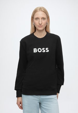 Lyshåret kvinde iført sølv øreringe, sort sweatshirt med "BOSS" i hvide bogstaver og lyseblå jeans mod en ensfarvet hvid baggrund.