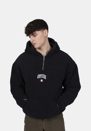 Black fleece pullover hoodie with zippered neck, front pocket, and "CRYSTAL" text with a small French flag. Soft texture, oversized fit.