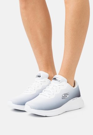 Persona che indossa scarpe sportive Skechers Skech-Lite bianche e grigie con lacci, in piedi su uno sfondo chiaro uniforme.