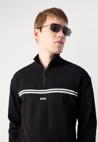 Μαύρος μισός ζιπ-πουλόβερ με υφασμάτινη υφή, λευκές οριζόντιες ρίγες και λογότυπο "BOSS" στο στήθος. Το μοντέλο φοράει σκούρα γυαλιά ηλίου.
