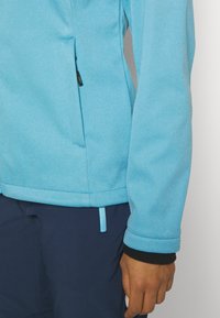Personne portant une veste bleu clair par-dessus un tee-shirt noir à manches longues et un pantalon bleu foncé, main détendue le long du corps sur un fond uni.