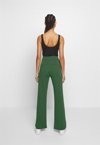 Vrouw met gevlochten haar, gekleed in een zwarte mouwloze top en hoge, groene wijde broek, staande voor een witte achtergrond.