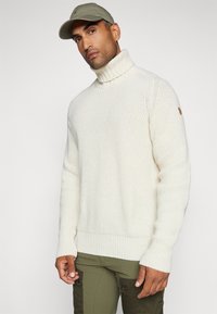 Maglione spesso a maglia di crema con colletto alto, orlo e polsini a costine. Indossato con pantaloni verde oliva, caratterizzati da un piccolo logo marrone sulla spalla.