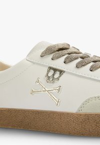 Weiße Leder-Sneaker mit einer strukturierten, hellbraunen Gummisohle. Mit metallischem Totenkopf- und Kreuzbones-Stickerei. Graue Schnürsenkel.