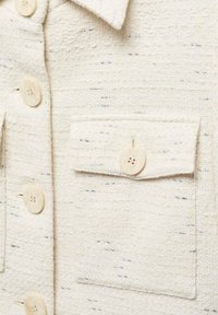 Kuvioitu kerma-valkoinen paita, joka on valmistettu kudotusta kankaasta. Paidassa on neljä pyöreää nappia, rintatasku ja hienovaraisia harmaita läikkiä pitkin pintaa.