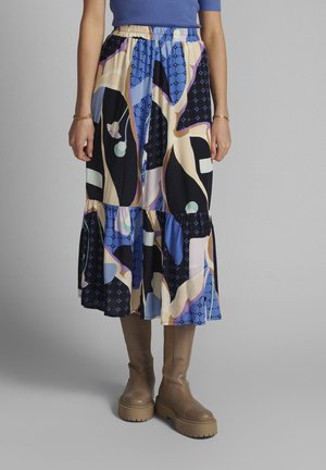 Vrouw draagt een midirok met een kleurrijk abstract patroon, een blauw bovenstuk en beige kniehoge laarzen, staande tegen een effen grijze achtergrond.