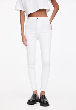 Slim-fit high-waist white jeans worn with a cropped white top and black slip-on chunky sole shoes on a plain background.