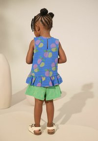 Blaues Oberteil mit buntem Obstmuster und Rüschensaum, kombiniert mit grün gestreiften Shorts. Kurze Ärmel und Knopfverschluss hinten.