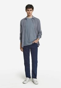 Ljusgrå skjorta med knappar över en ljusblå t-shirt, kombinerat med mörkblå jeans och vita sneakers, vilket ger en avslappnad lager-på-lager-look.