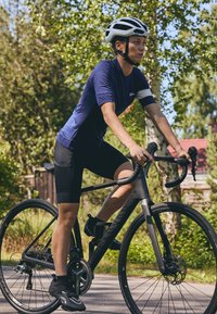 Juodas kelių dviratis su elegantišku rėmu, juodais padangomis ir Shimano komponentais. Dviratininkas vilki mėlyną marškinėlį, juodas šortus ir juodus dviratininkų batus.