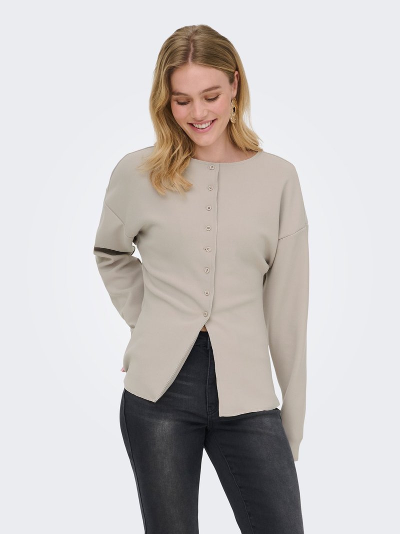 Donna sorridente con capelli biondi alla spalla, indossa una maglia beige a maniche lunghe con bottoni e jeans scuri, in piedi con una mano dietro la schiena.