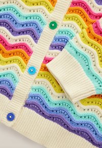 Cardigan tricoté avec une base crème et des rayures ondulées multicolores en violet, bleu, vert, orange, rose et jaune. Grands boutons bleus.