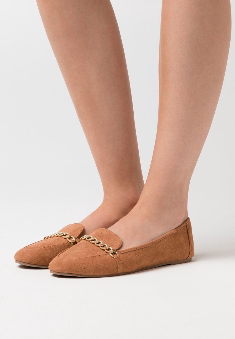 Pieds portant des chaussures plates en daim marron avec un détail chaîne dorée sur le devant, sur un fond clair uni.