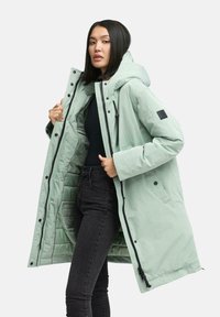 Mintgrüner Parka mit Kapuze, ausgestattet mit schwarzen Akzenten, Knopfverschlüssen und einem inneren gefütterten Futter, getragen über schwarzen Jeans.