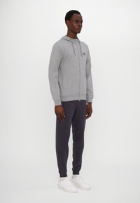 Sivi zip-up hoodie s kapuljačom, u kombinaciji s tamnosivim hlačama. Bijele tenisice upotpunjuju odjevnu kombinaciju, s jednostavnim dizajnom i ravnim krojem.