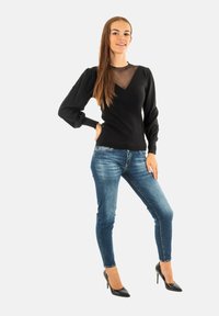 Maglia nera a maniche lunghe con scollatura a V e dettaglio in tessuto trasparente; abbinata a jeans skinny blu e tacchi alti neri.