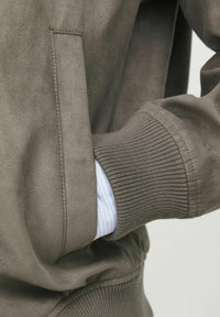 Jachetă din suede maro cu manșete ribbed, având un buzunar lateral. Materialul cămășii de culoare albastru deschis este vizibil pe dedesubt. Textură netedă și design simplu.