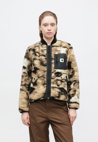 Chaqueta bomber de pelaje sintético en beige con manchas negras, con cremallera frontal, bolsillo negro y puños acanalados. Textura suave y ajuste relajado.