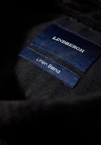 Zwarte stof met blauwe labeltags met de teksten "LINDBERGH" en "Linnenmix." De labels hebben een gladde textuur en gestikte randen.