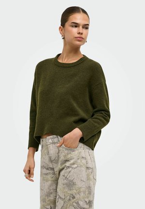 Femme aux cheveux attachés porte un pull vert olive et un pantalon gris clair à motifs, debout avec une main dans la poche sur un fond uni.