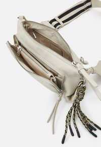 Helle beige Leder-Umhängetasche mit zwei Reißverschlussfächern, texturiertem Finish und einem mehrfarbigen geflochtenen Quasten-Detail.