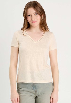 Junge Frau mit hellbraunen Haaren trägt ein beigefarbenes, kurzärmeliges V-Ausschnitt-Top und hellblaue Jeans, steht vor einem schlichten weißen Hintergrund.