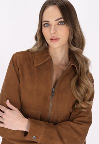Giacca in suede marrone con zip frontale, colletto e due tasche. Tessuto liscio, vestibilità rilassata e dettaglio di cucitura sottile sulle maniche.