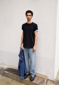 T-shirt noire à manches retroussées, jean bleu clair ample et sabots noirs. Une veste en denim bleu est tenue dans la main gauche contre un fond neutre.