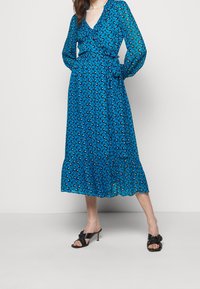 Vestido largo azul con estampado floral, mangas largas, escote en V, detalles con volantes y un dobladillo en capas. Modelo combinado con tacones negros abiertos por delante.