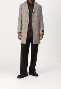 Abrigo de patrón de espiga en beige y marrón, con dos bolsillos frontales y cierre de tres botones, combinado con camisa y pantalones marrones oscuros.