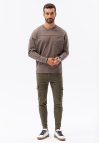 Hnědá košile s dlouhým rukávem a kapsou na hrudi, ve spojení se zelenými krajkovými kalhotami a bílými teniskami s černými tkaničkami a hnědými podešvemi.