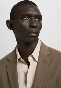 Hellbeiges Hemd unter einem hellbraunen Blazer mit Stegkragen. Glatter Stoff mit feiner Textur. Nahaufnahme eines Mannes.