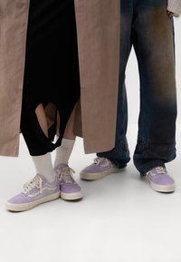 Ljust lila sneakers med vita sulor, beige överdimensionerad kappa, svart plagg med utskärning och lösa blå jeans. Texturerade och lager-på-lager stilar.