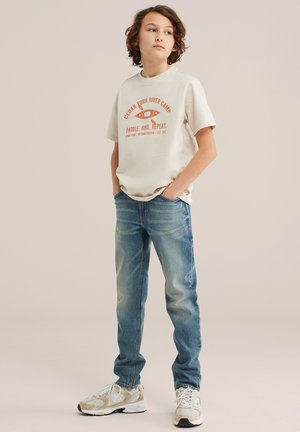 Otrok s kodrastimi lasmi, oblečen v bež majico Cedar Fork River Camp, modre kavbojke in bele superge, stoji z rokami v žepih.