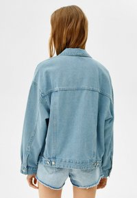 Světle modrá denimová bunda s uvolněným střihem, klasickým límcem, zapínáním na knoflíky a viditelným švem na zadní straně.
