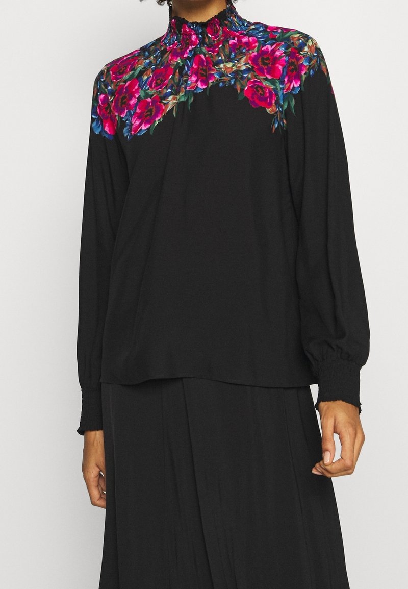 Femme portant un haut à manches longues noir avec des motifs floraux roses vifs et bleus autour du col haut et des épaules.