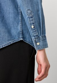 Denim overhemd met lange mouwen, lichtblauwe kleur, knoopmanchetten en een licht gebogen onderkant. Draagt over zwarte broeken.