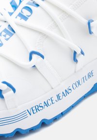 Λευκό αθλητικό παπούτσι με μπλε λεπτομέρειες, διαθέτοντας υφασμάτινο επάνω μέρος με υφή, ελαστικά κορδόνια και το λογότυπο "VERSACE JEANS COUTURE" τυπωμένο στη σόλα.