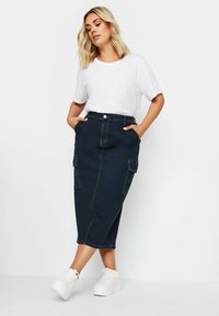 Ciemnoniebieska spódnica midi z przodu z kieszeniami, wysokim pasem i prostym krojem, zestawiona z białą koszulką i masywnymi białymi butami.