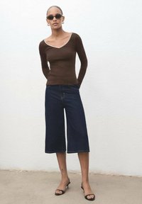 Rjava dolga majica z dolgimi rokavi in belim ovratnikom, v kombinaciji s temno denim širokimi culottes hlačami in črnimi sandali na peto. Kratki lasje in sončna očala.