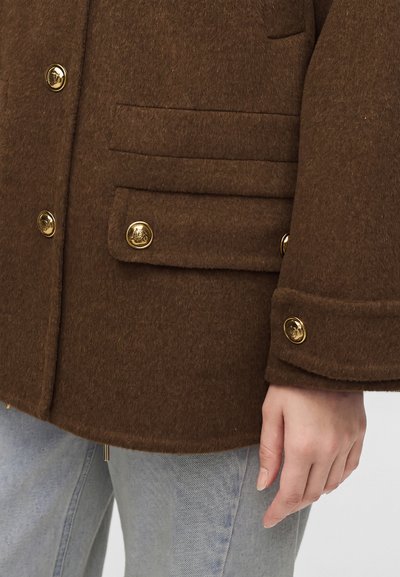 Blazer en laine marron avec boutons dorés, poche à rabat et détails aux poignets porté sur un jean bleu clair, montrant une main sur le côté.