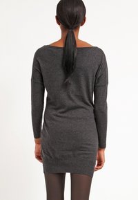 Femme avec une longue queue de cheval noire portant une robe-pull grise foncée à manches longues et des collants noirs, debout de dos sur un fond blanc.