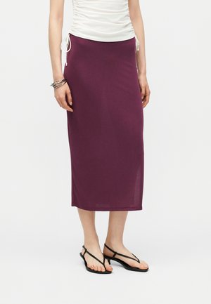 Femme portant une jupe midi couleur prune, un top blanc froncé, des sandales noires à lanières et des bracelets argentés au poignet gauche.